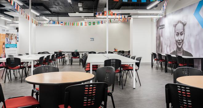 A large seating area with a combination of long, rectangular tables and small circle tables. Chairs surround the tables. Different countries' flags hang from the ceiling. Victoria University banner hangs in the background.