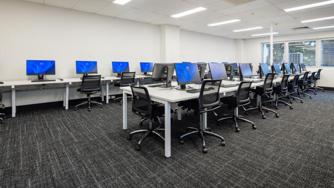 A room with computers. In the middle of the room is a long table with computers on it and chairs surrounding it. At the far wall is another table with computers and chairs.