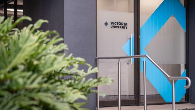 A door with the sign "Victoria University" engraved on it. A plant is in the foreground, out of focus.