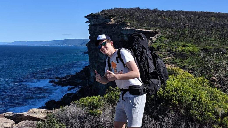 Denis giving a thumbs up in front of a spectacular view - water to the left and a cliff to the right.