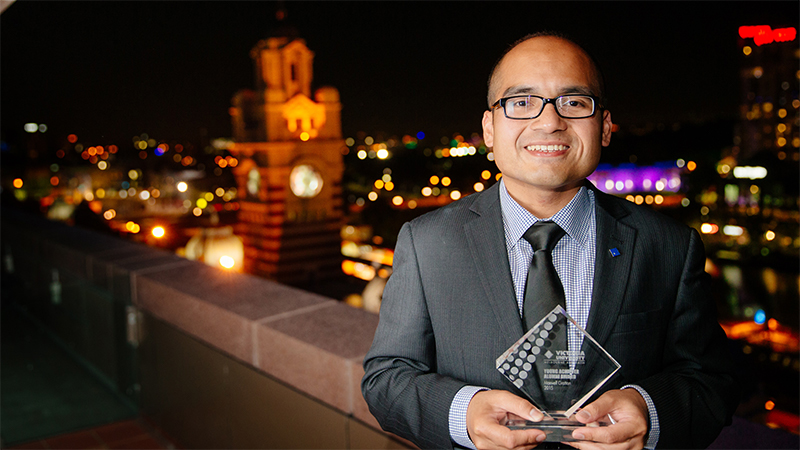 Maxwell Gratton holding his award, at night with a view of Flinders Street station behind him.