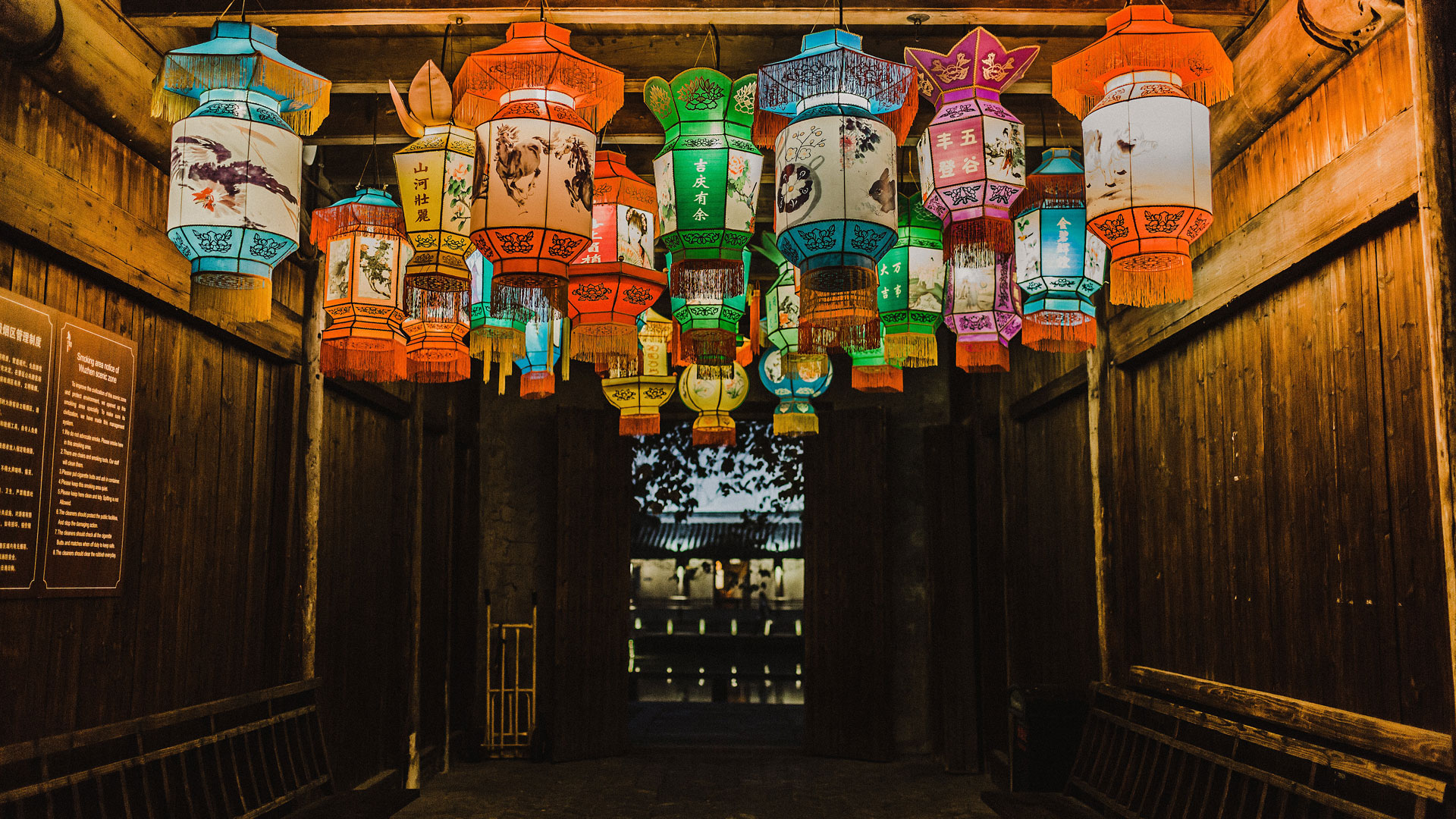 Paper balloons light up the roof of a wooden hallway with doors open to show another building in the distance.