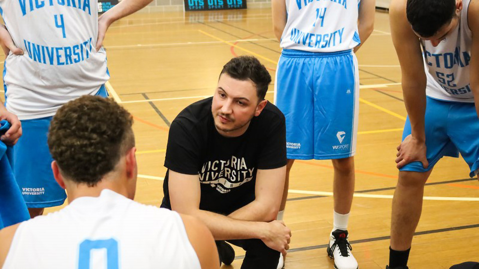 A coach kneeling down on a basketball court, surrounded by 4 people standing up in basketball uniforms. The coach is looking at one of the players.