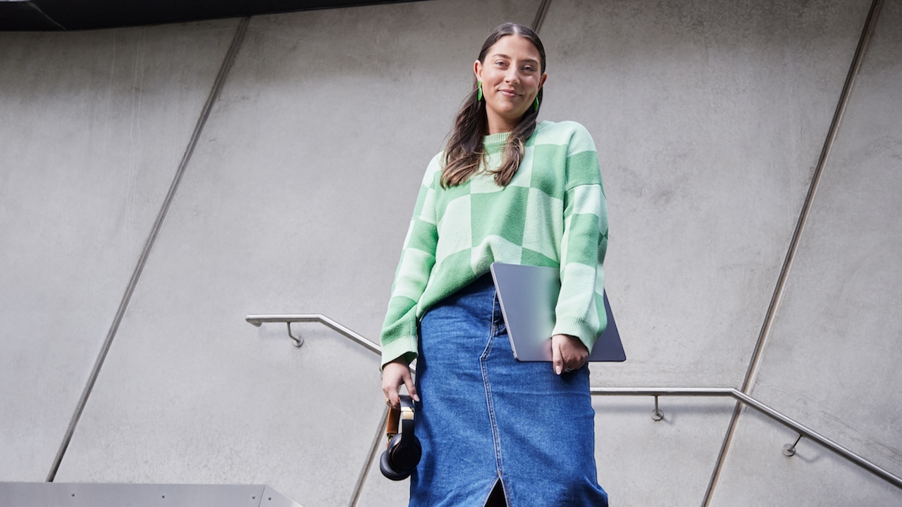 Alexandra, wearing a green checkered sweater, smiles while leaning against a wall. She is quoted saying she chose VU because of the Block Model.