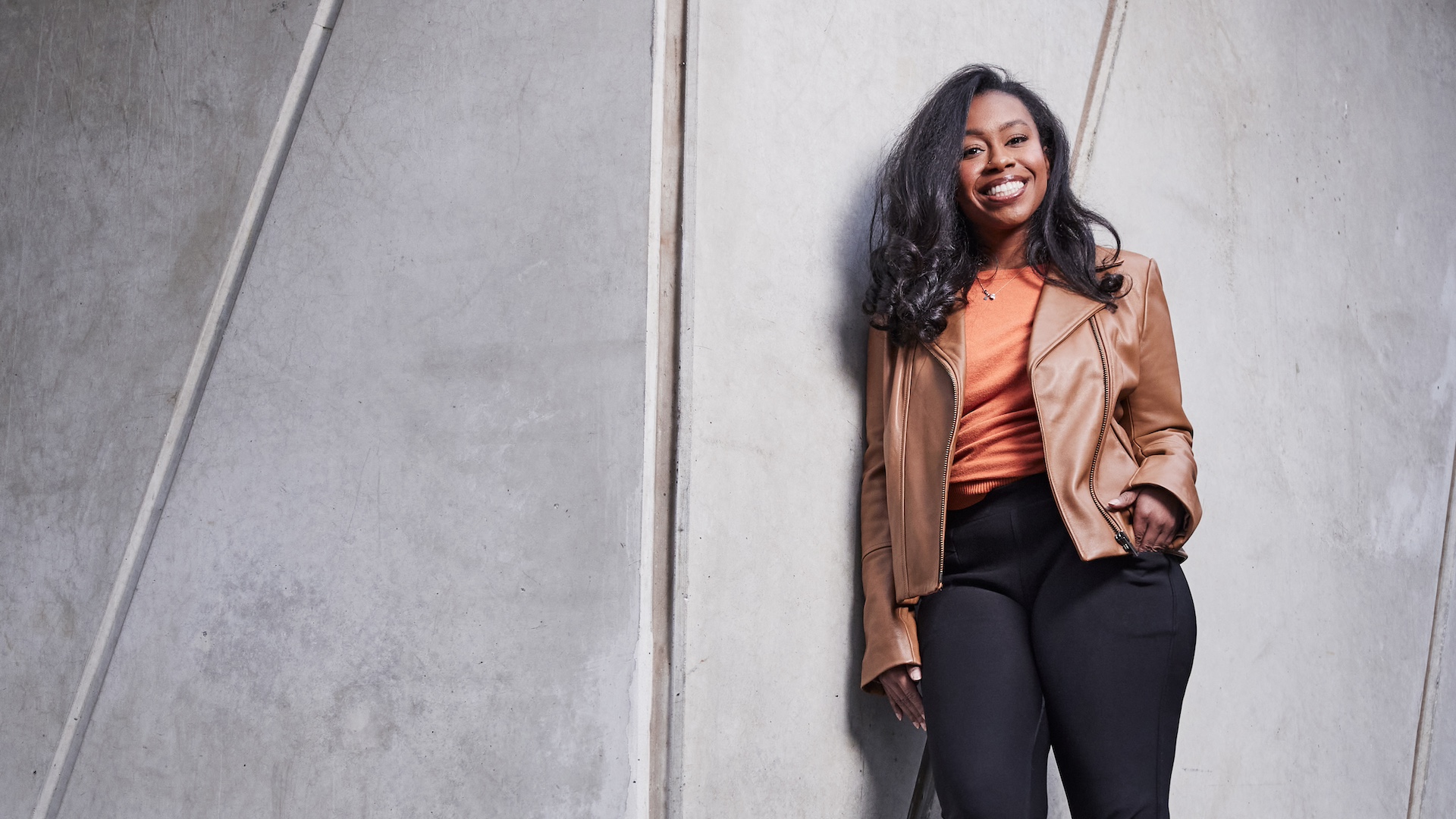 Shelbey, wearing a tan jacket and orange shirt, smiles while leaning against a concrete wall. She states she chose VU for its diversity and support.