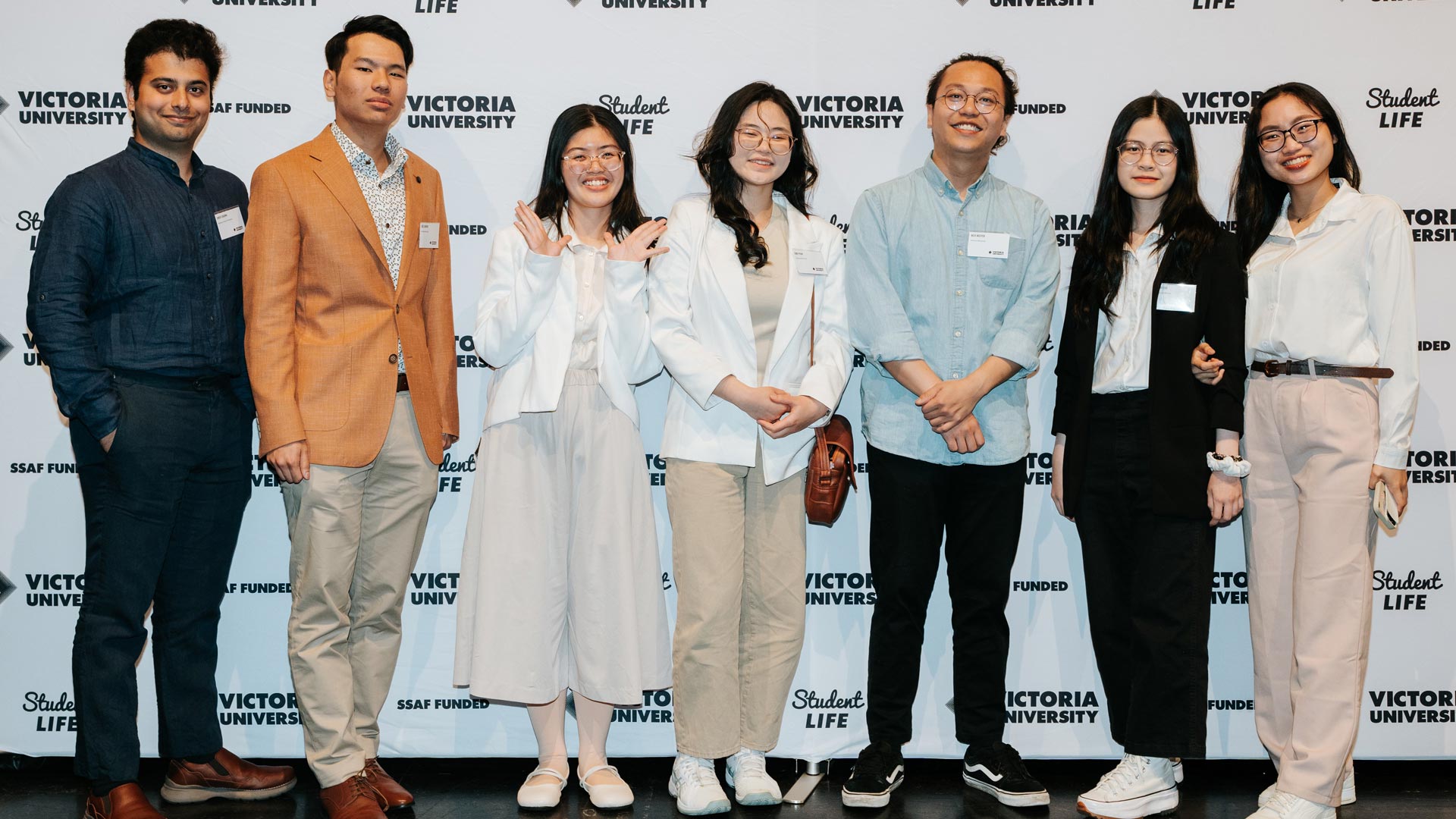 7 students in formal wear standing in front of a VU backdrop, smiling for camera.