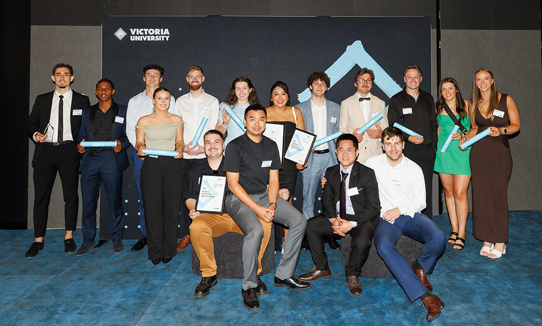 A group of young adults in formal dress pose with awards in front of a Victoria University banner