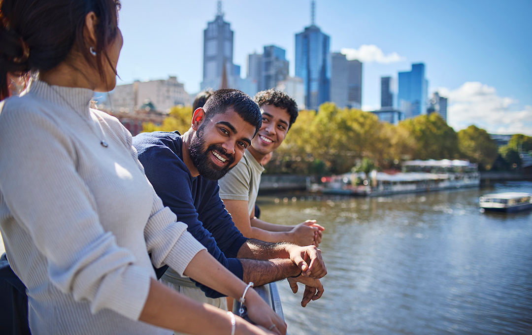A group of international students smile on a bridge over a wide river, in front of a modern city skyline