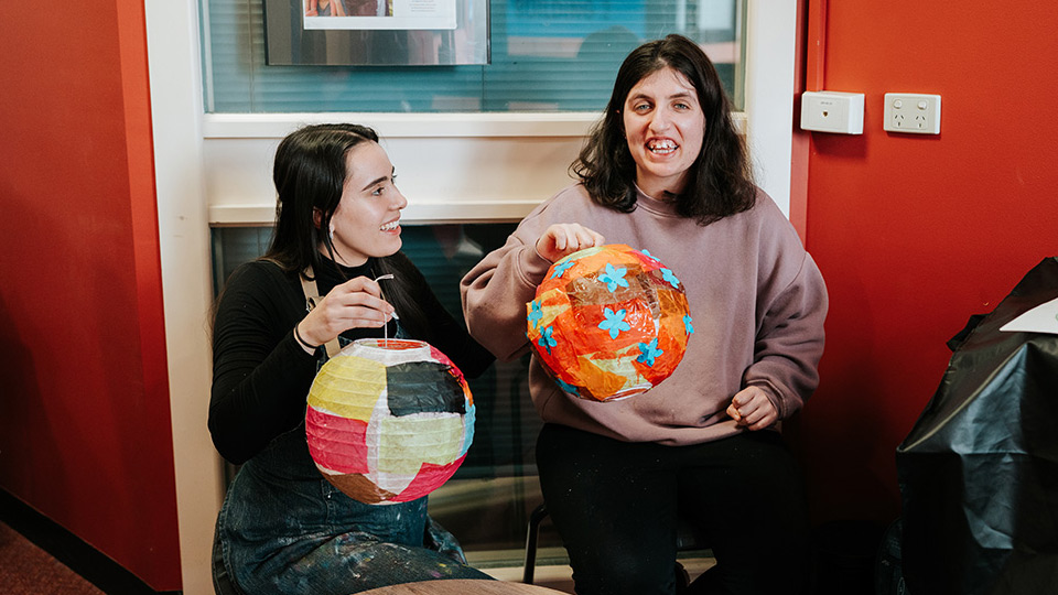 Two young women are smiling and displaying handmade lanterns. Bernie is on the right hand side, wearing a light purple jumper.