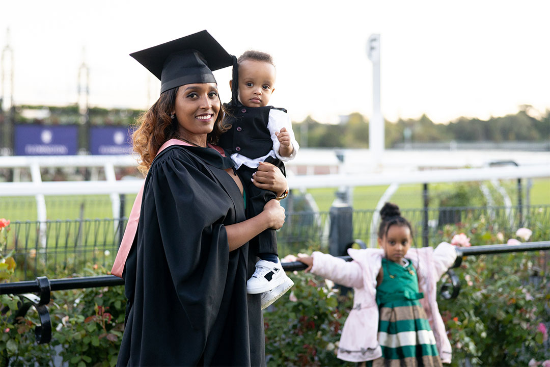 Woman in a graduation hat and gown holds a child at Flemington Racecourse. Another child stands behind her, holding on to a nearby fence.