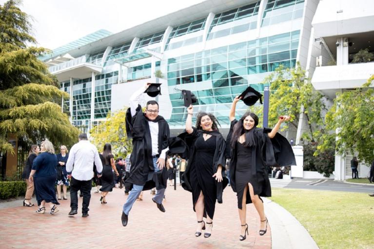 three graduates jumping for joy, holding their caps, outside the Flemington Racecourse