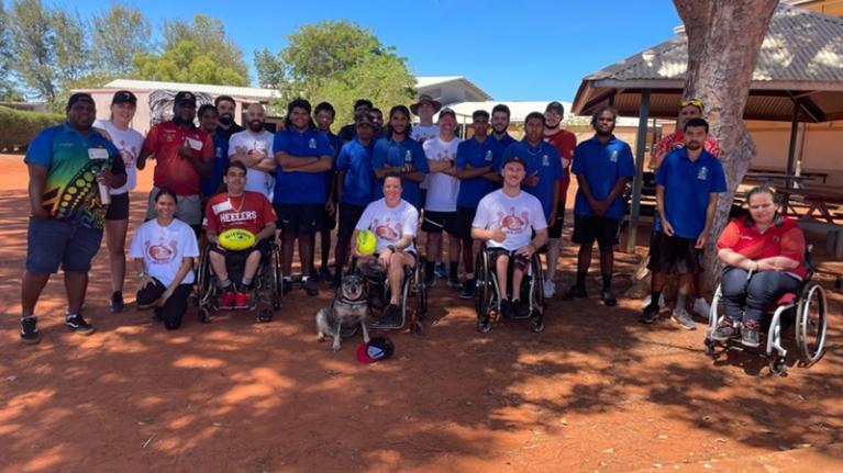 Group of people, some Indigenous, some in wheelchairs, on dry, red earth outside a building