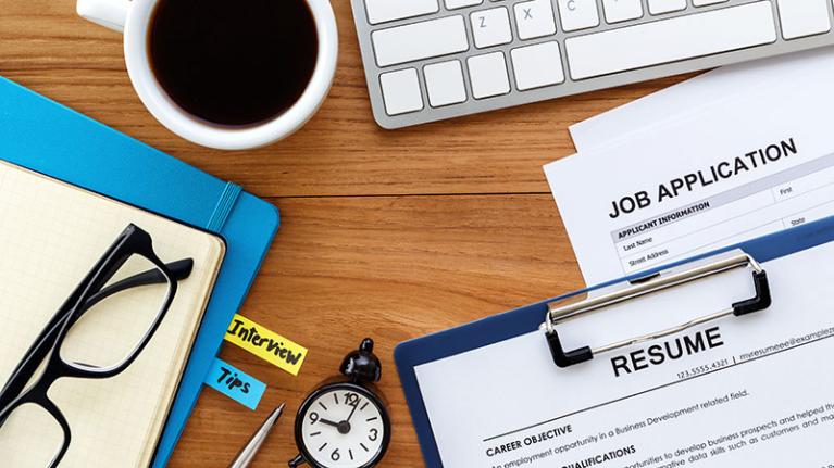 A desk with a resume, job applications, laptop, glasses and coffee on its surface.