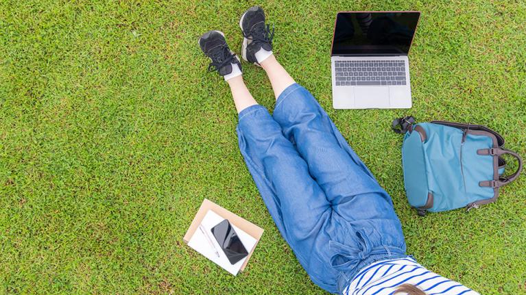 A student sitting on grass with laptop, notrbooks, phone and bag spread out around them.