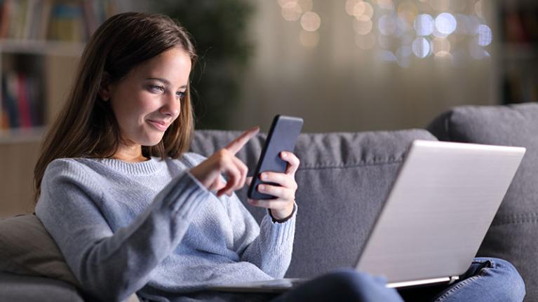 Student at home holding phone with laptop on lap.