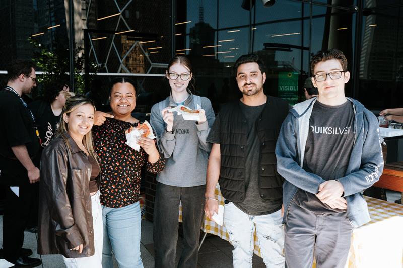 A group of young adults smile in the sun in front of a modern glass window