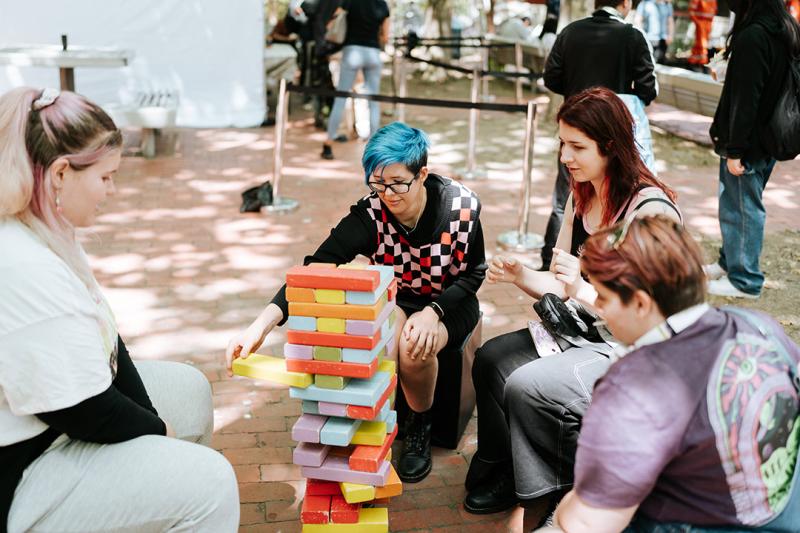 A group of students with bright hair play a block-stacking game in a sunny courtyard