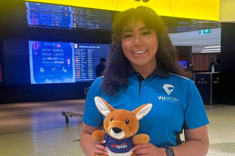 Stella Rose Lay standing in an airport wearing a VU Sport shirt holding a plush kangaroo with the UniSport logo.
