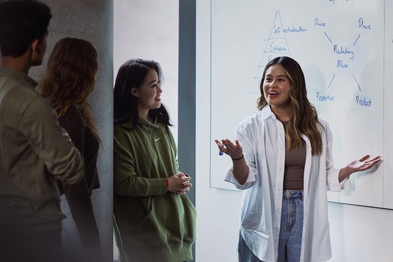 A female student presenting to 3 students. Behind her is a whiteboard with diagrams about marketing principles.