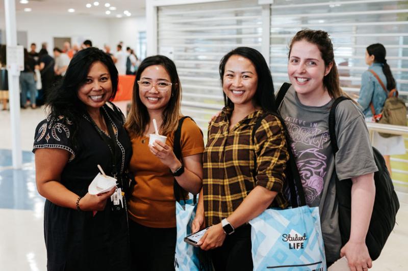 Four students indoors smiling at the camera. Two students are holding small ice cream cups. One student is wearing a Student Life tote bag.