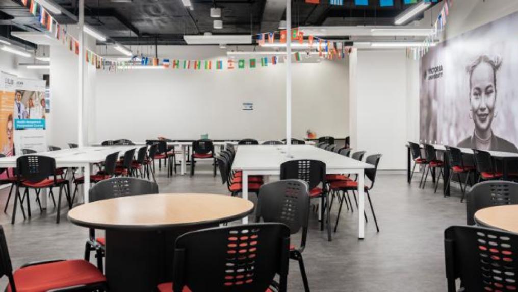 A large seating area with a combination of long, rectangular tables and small circle tables. Chairs surround the tables. Different countries' flags hang from the ceiling. Victoria University banner hangs in the background.