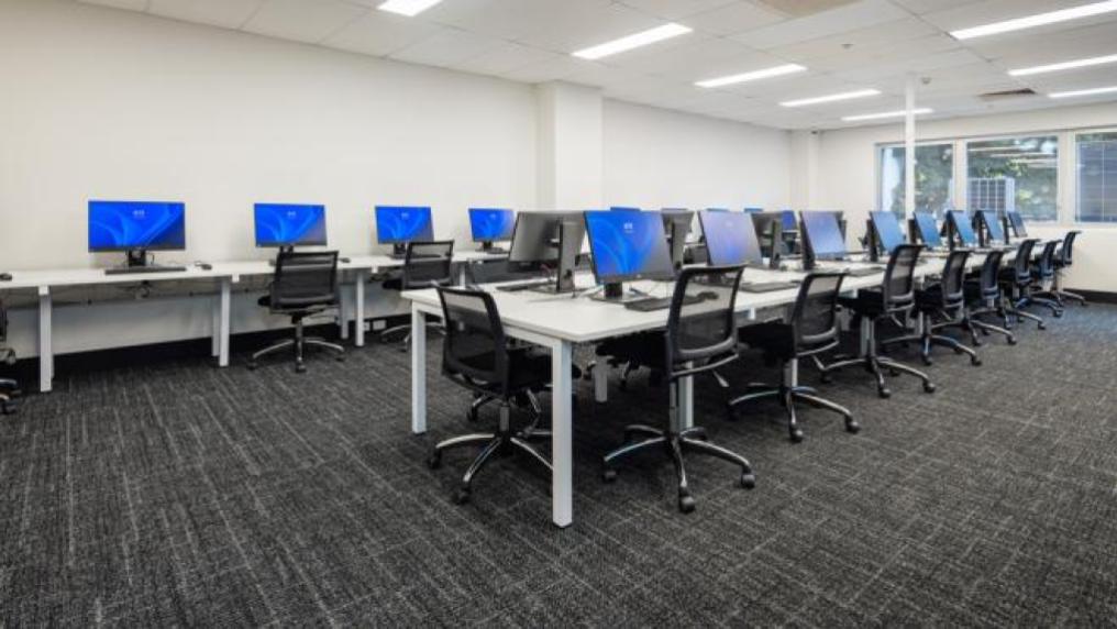 A room with computers. In the middle of the room is a long table with computers on it and chairs surrounding it. At the far wall is another table with computers and chairs.
