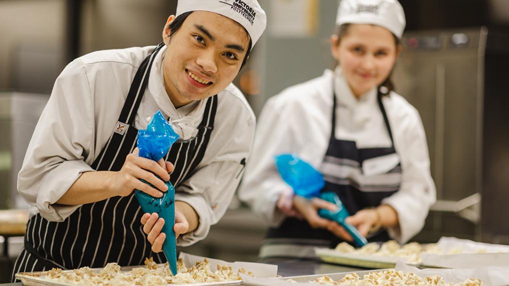 A young man and woman in chef uniforms smile while adding sauce to a platter