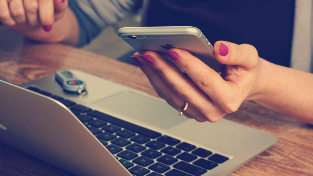 A hand holds a phone while hovering over a laptop