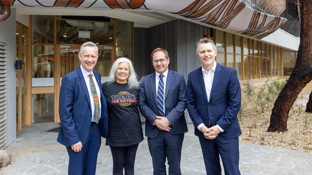 Three men in suits stand with a woman wearing a Treaty t-shirt in front of an Indigenous design over a doorway