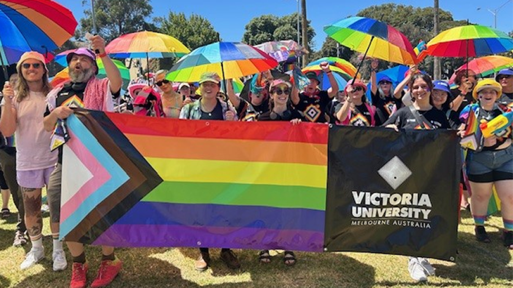 A group of more than 40 people march behind a Pride Flag, in gardens in St Kilda. It is a sunny day and the marchers look happy, wearing rainbow colours and carrying rainbow parasols.