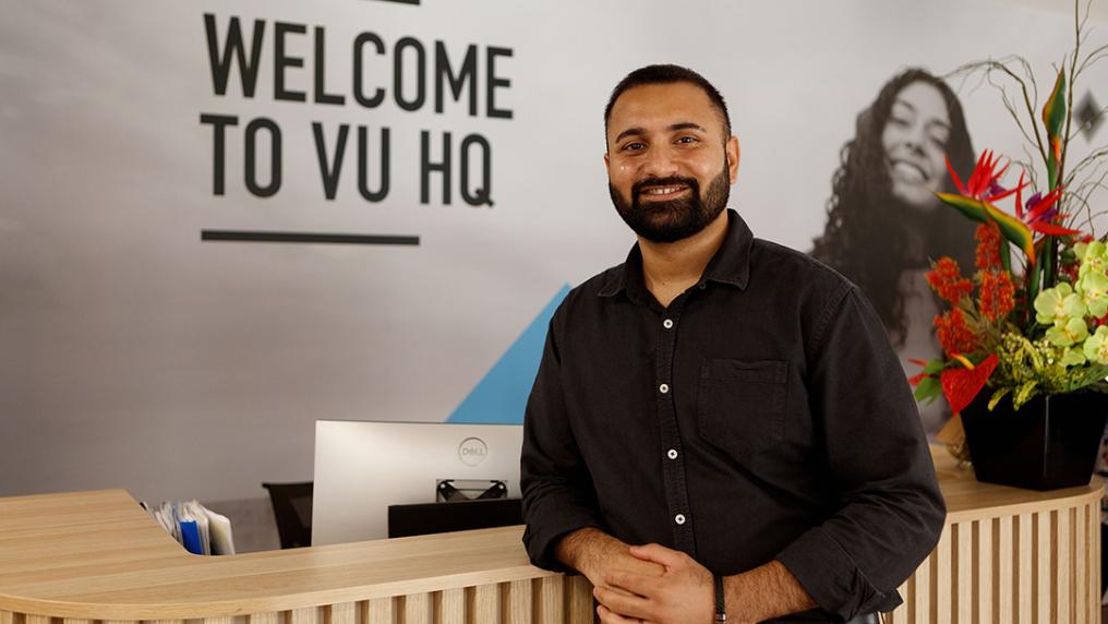 A smiling man leans on a reception desk. On the wall behind is written 'Welcome to VU HQ'
