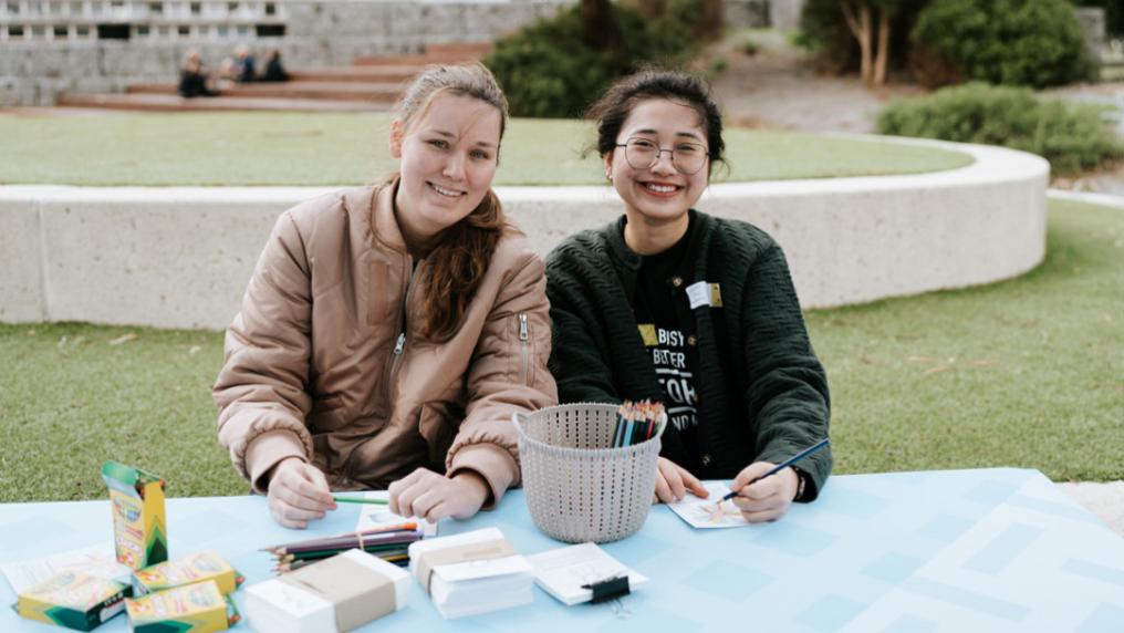 Two students sitting behind a table outside. On the table are some art supplies, including crayons, pencils and paper.