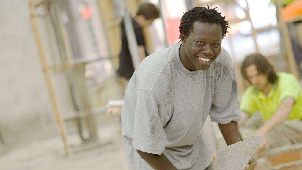A young man holds a trowl and smiles while laying bricks in a large workspace
