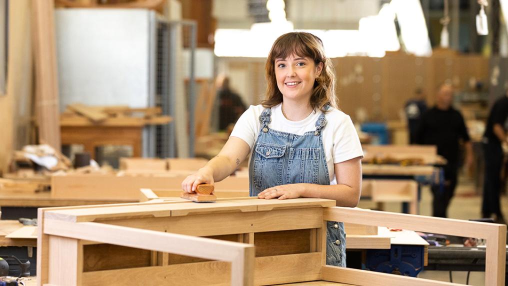 A young woman in dungerees smiles while sanding wooden furniture in a large workshop