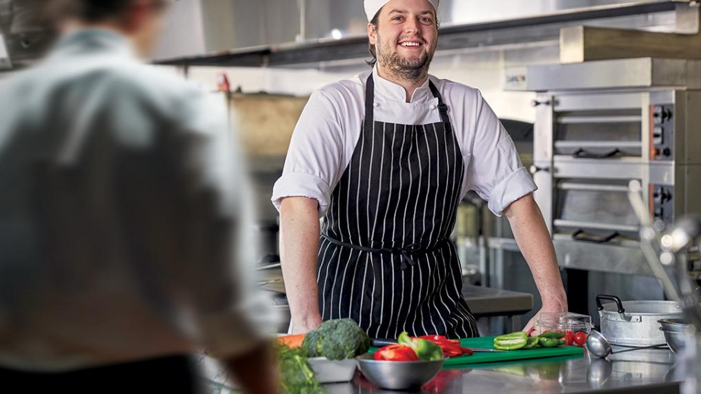 A young man in a chef uniform beams while preparing food in a professional kitchen