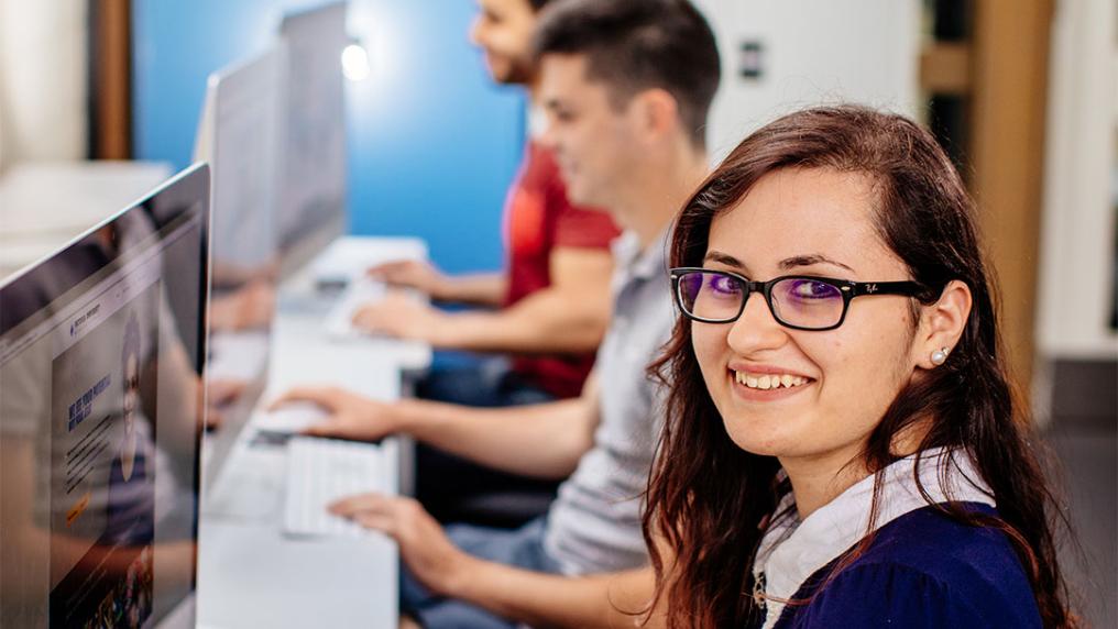 Student in glasses sitting at a desktop Mac, smiling at the camera.