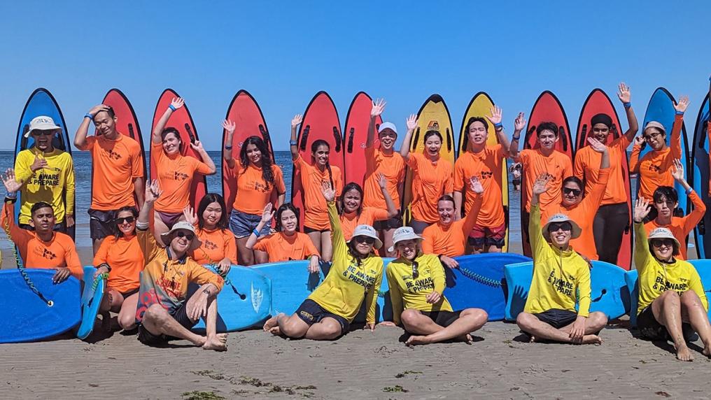 VU students in brightly-coloured swimgear sitting on the beach and waving at the camera.