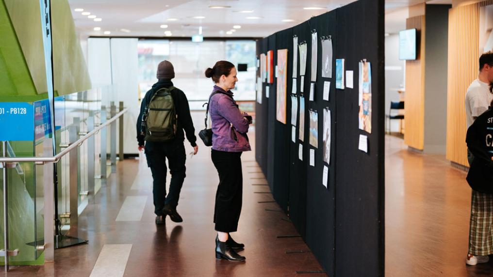 A woman looking at art displays on a temporary wall. The wall is set in the library corridor, with people walking in the background.