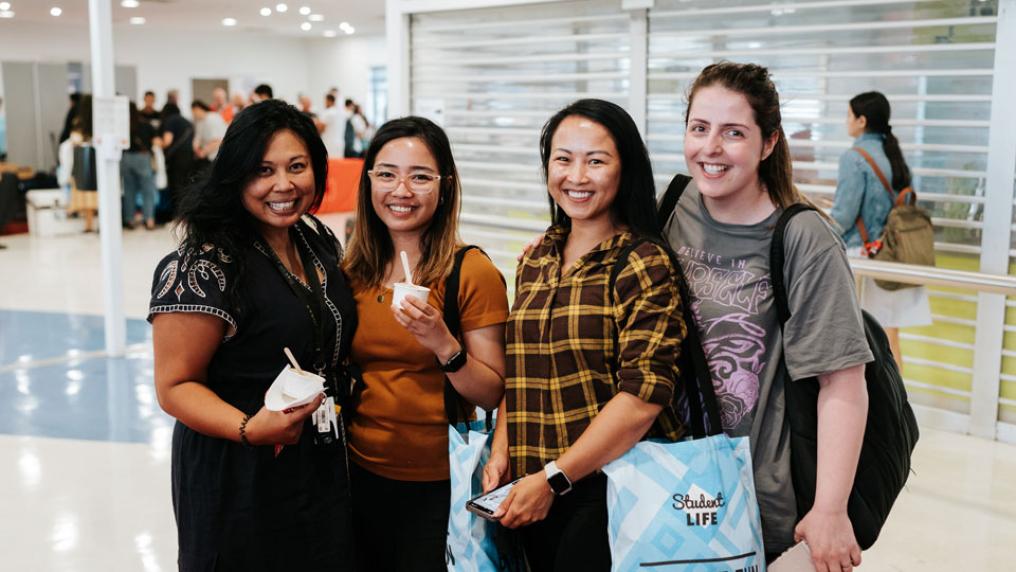 Four students indoors smiling at the camera. Two students are holding small ice cream cups. One student is wearing a Student Life tote bag.