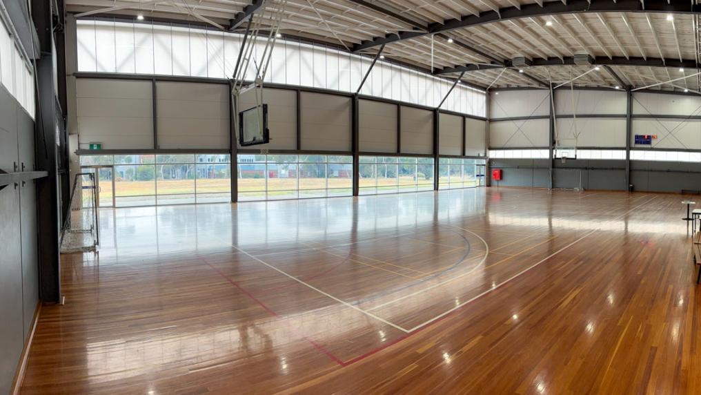 A large indoor court with a basketball hoop on either side and a digital scoring board. One wall has windows that look out onto nearby grass and buildings.