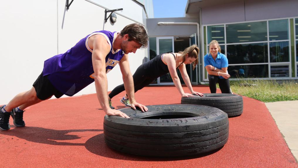 Two people outside in a push-up postion, with their hands on top of a tyre. A personal trainer is leaning and observing them in the background.