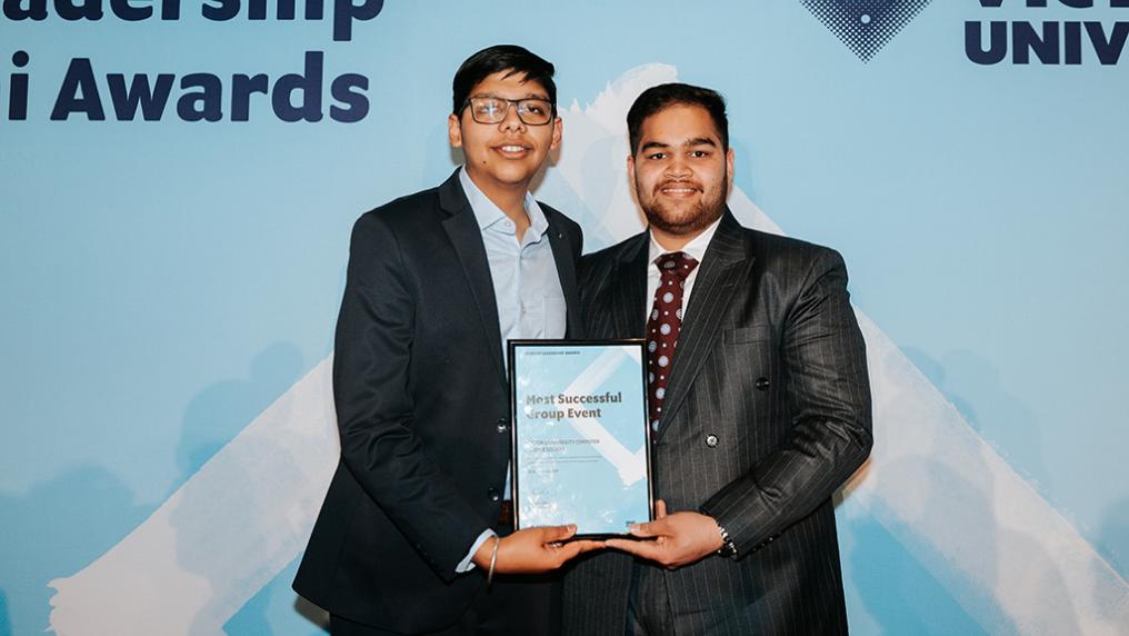 Two young men in smart suits pose with an award in front of a Victoria University banner