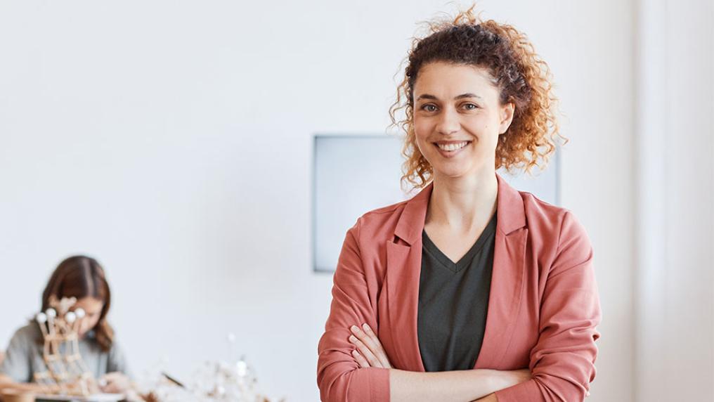 A woman in a red blazer with curly hair stands in front of a group of primary school-aged kids in a classroom setting.