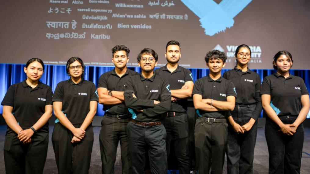 A multicultural group of friendly young adults in black staff uniform in front of a welcome banner with different languages and Victoria University branding
