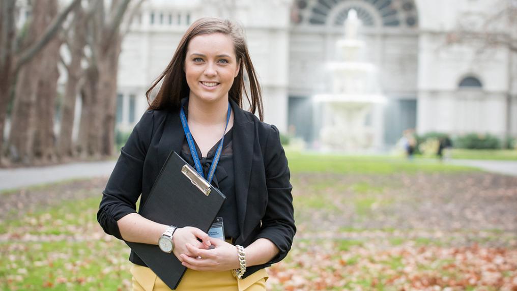 A young woman with a clipboard strids across and autumn lawn in front of an elegant building