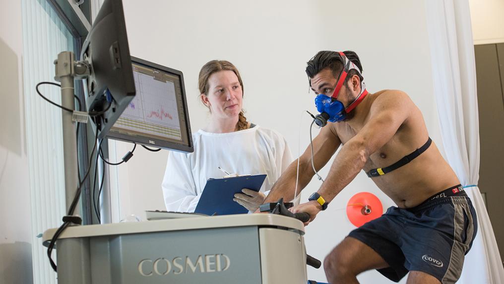 man hooked up to monitoring equipment rides an exercise bike, watched by a woman in a lab coat