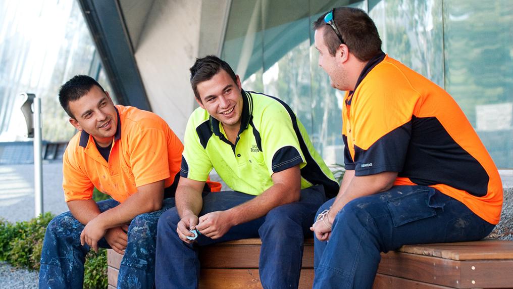 three young men in trades uniforms chat outside a modern campus building