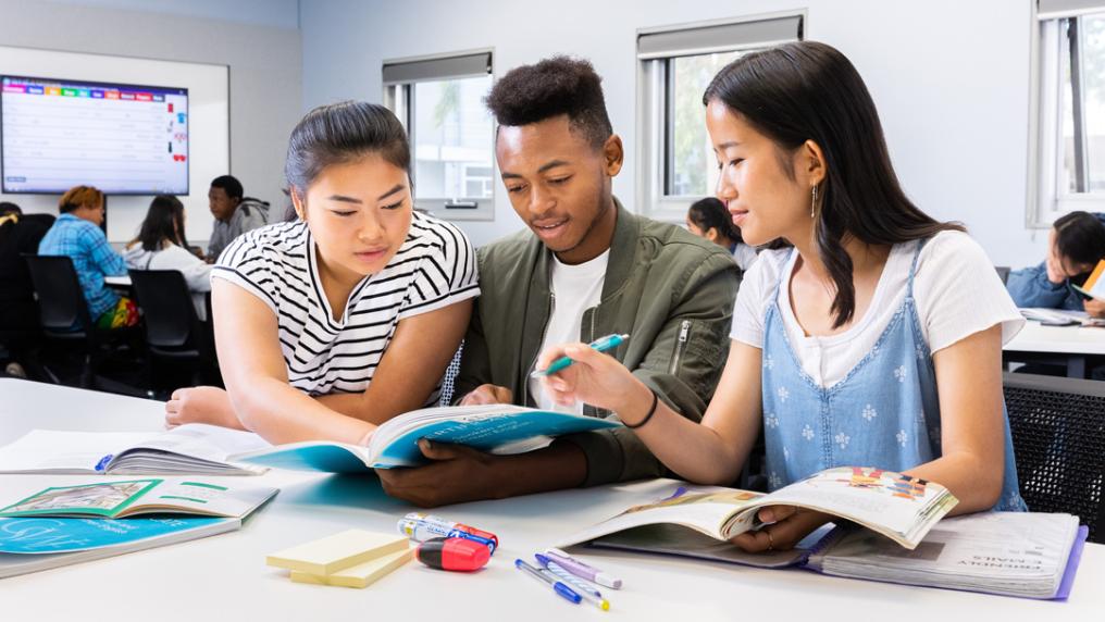 Students in a classroom. Three students in the foreground are sitting down and looking at the same book.