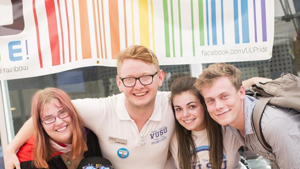 two male and two female students under a rainbow banner, at an orientation stall