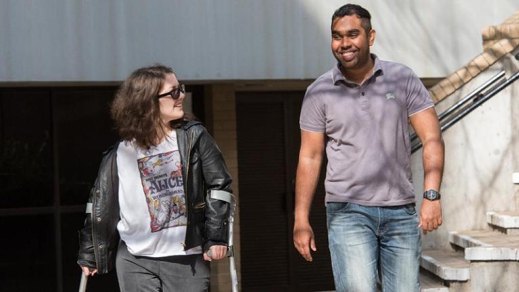 Two people walking and smiling. One of them is using crutches.